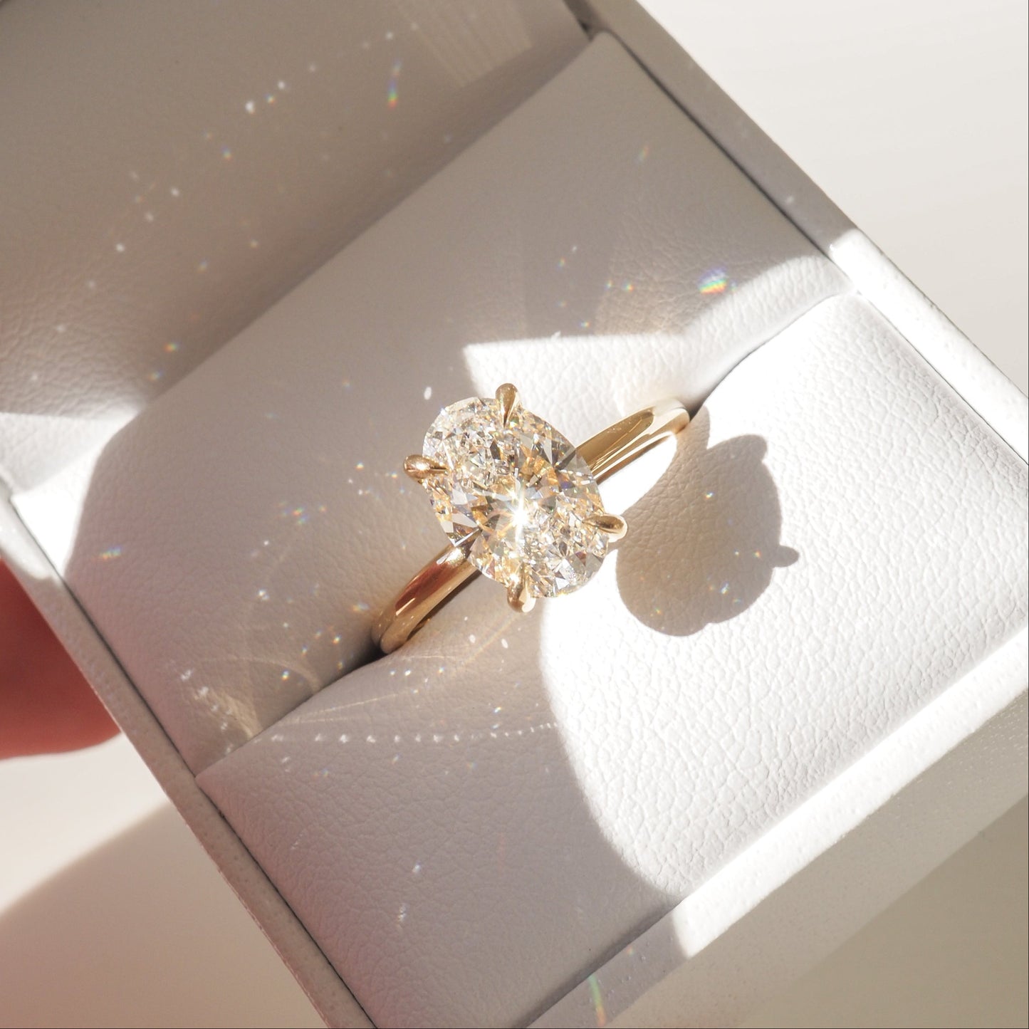 A stunning oval diamond ring in a gold setting sits in an elegant white jewellery box, illuminated by sunlight, casting sparkling reflections.