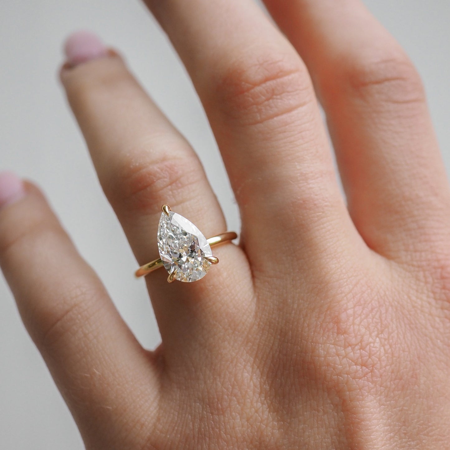 Close-up of a hand wearing a gold ring with a large, pear-shaped diamond. The backdrop is a soft, neutral tone, emphasising the jewellery.