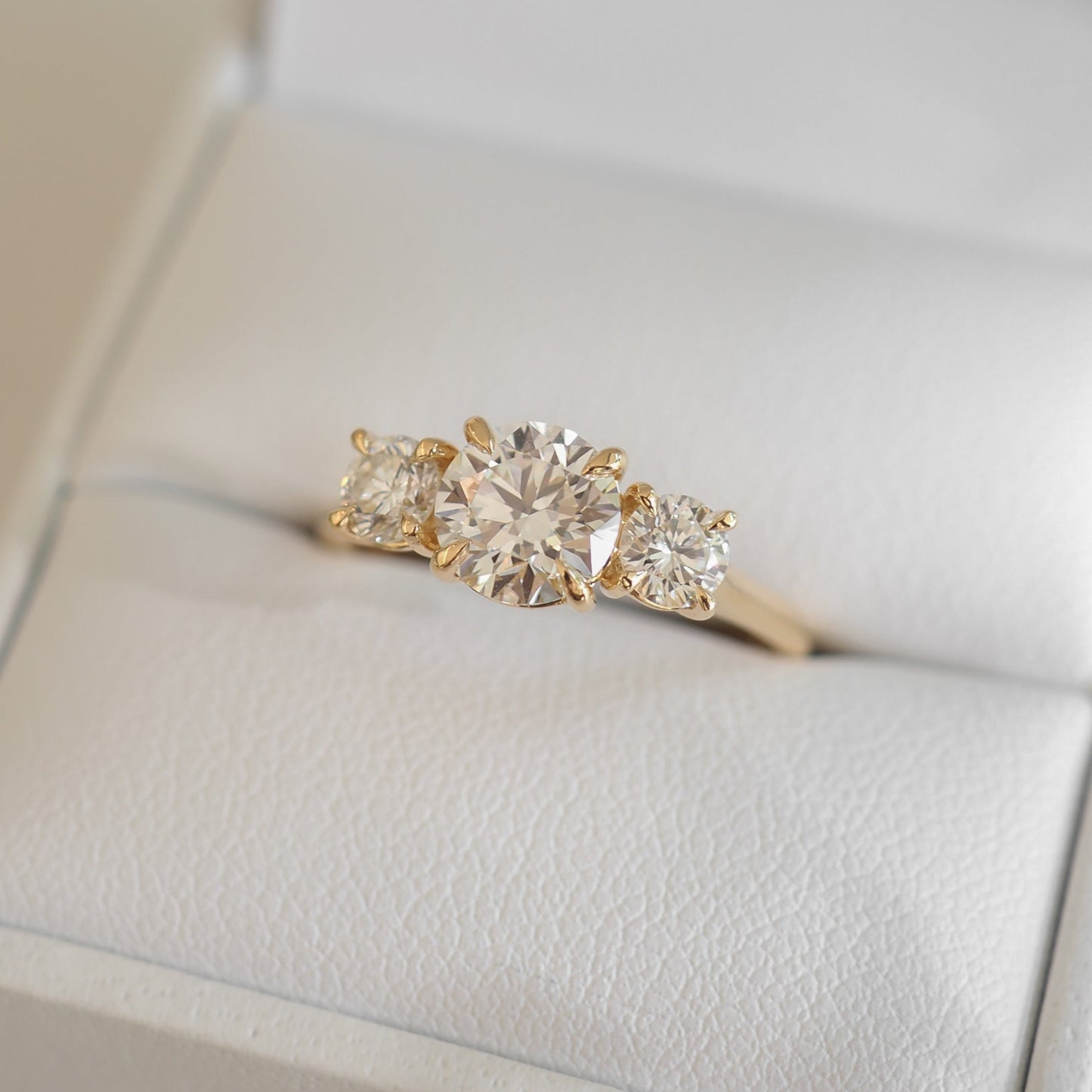 A gold engagement ring with a large round diamond flanked by two smaller square diamonds is displayed in a white jewellery box.