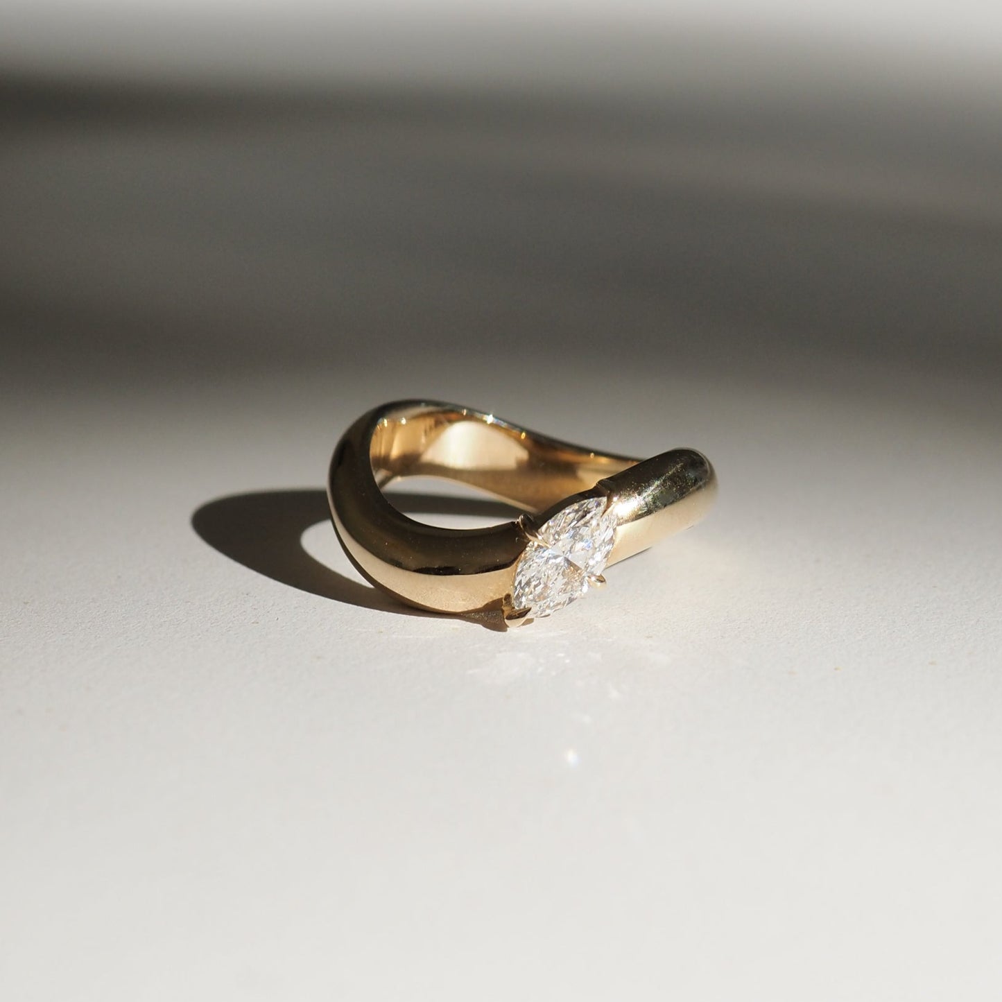 Gold ring with a marquise diamond set in a unique twist design, casting a soft shadow on a neutral surface. Luxurious and elegant jewellery.