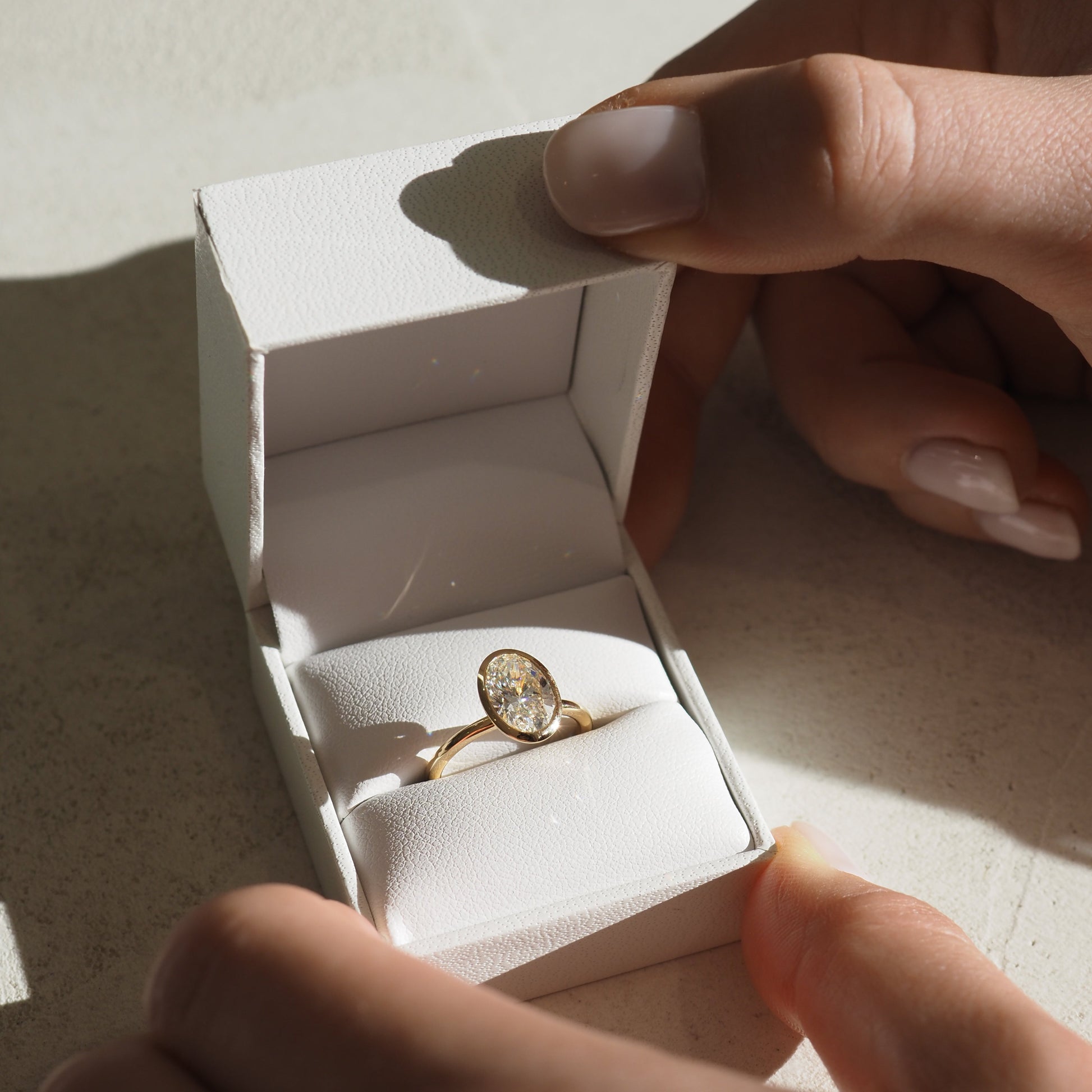 Hands hold an open white jewellery box with a gold ring featuring a large oval diamond. Sunlight casts soft shadows, highlighting the elegant design.