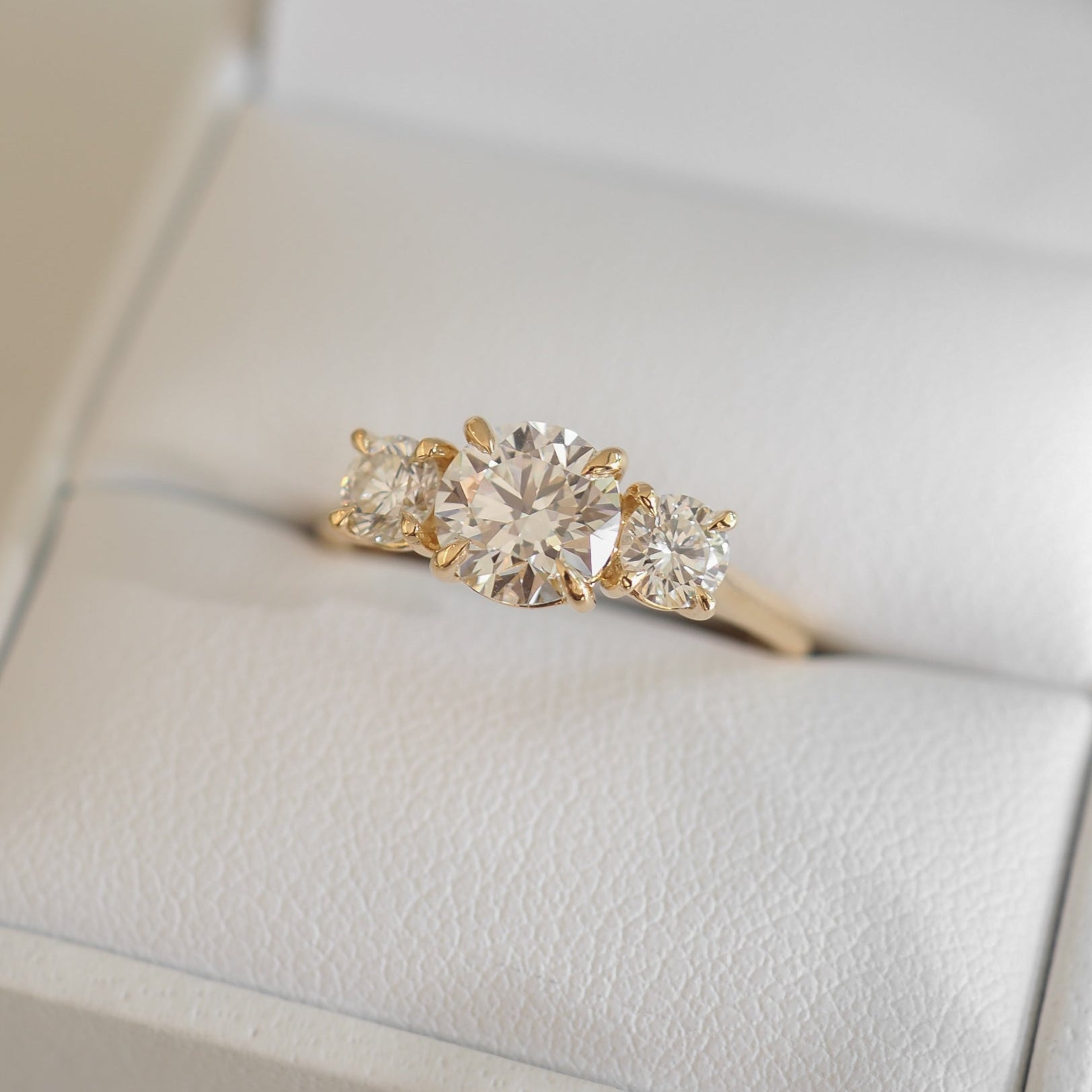 A gold engagement ring with a large round diamond flanked by two smaller square diamonds is displayed in a white jewellery box.